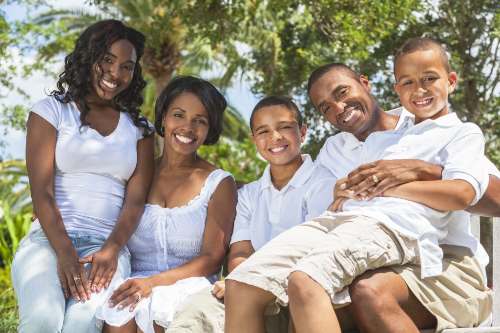 A happy black African American family of two parents and three children, two boys one girl, sitting together outside.