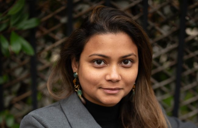 Noémie Gaddarkhan, assise sur un banc, en veste grise, regardant l’objectif avec assurance, devant une grille végétalisée.