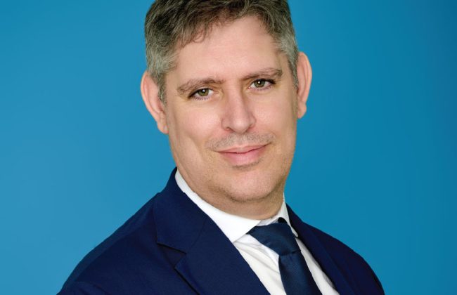 Portrait de Frédéric Guyonnet en costume bleu marine, tendant la main en avant, sur fond bleu uni.