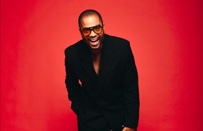 Ludovic Louis, trompettiste martiniquais, en costume noir et baskets blanches, souriant, tenant sa trompette sur fond rouge vibrant.