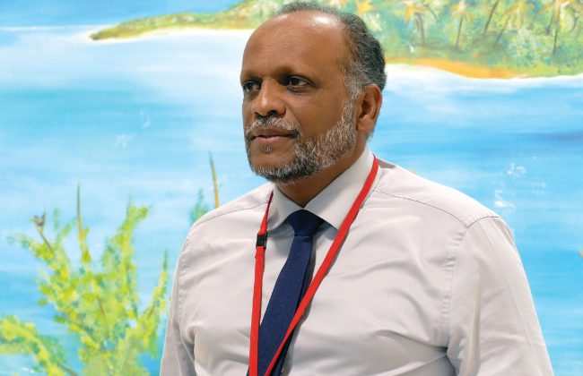 Patrice Phoudiah, directeur commercial Guadeloupe et Îles du Nord de la Caisse d’Épargne CEPAC, en chemise blanche et cravate bleue, devant une fresque tropicale