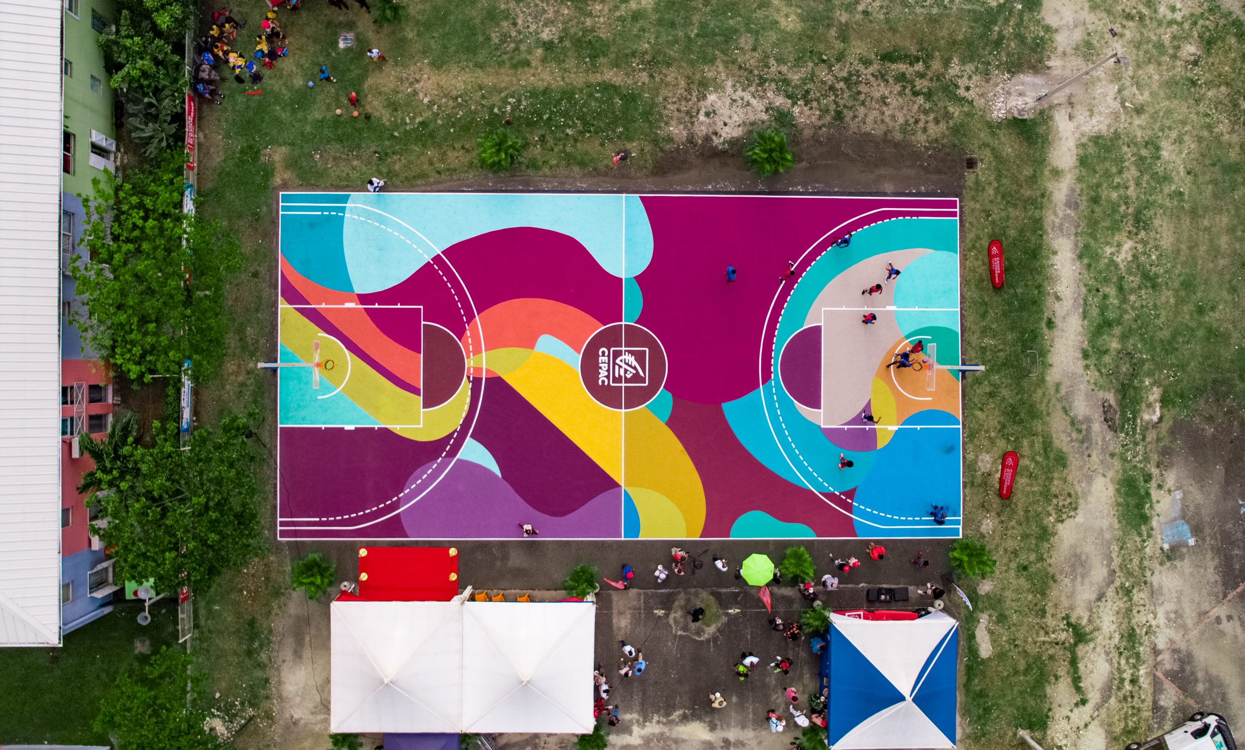 Vue aérienne d’un terrain de basket 3x3 aux couleurs vives, avec des personnes autour et des tentes installées en périphérie