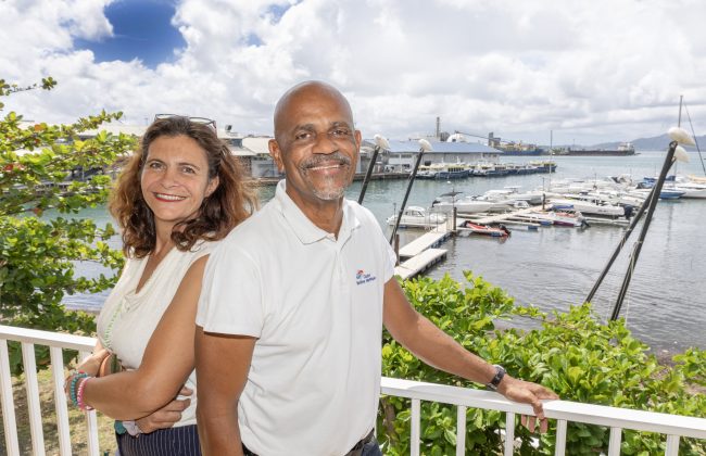 Alexandrine Milesi et Emmanuel Lise posent devant une marina en Martinique, souriants, sous un ciel partiellement nuageux.