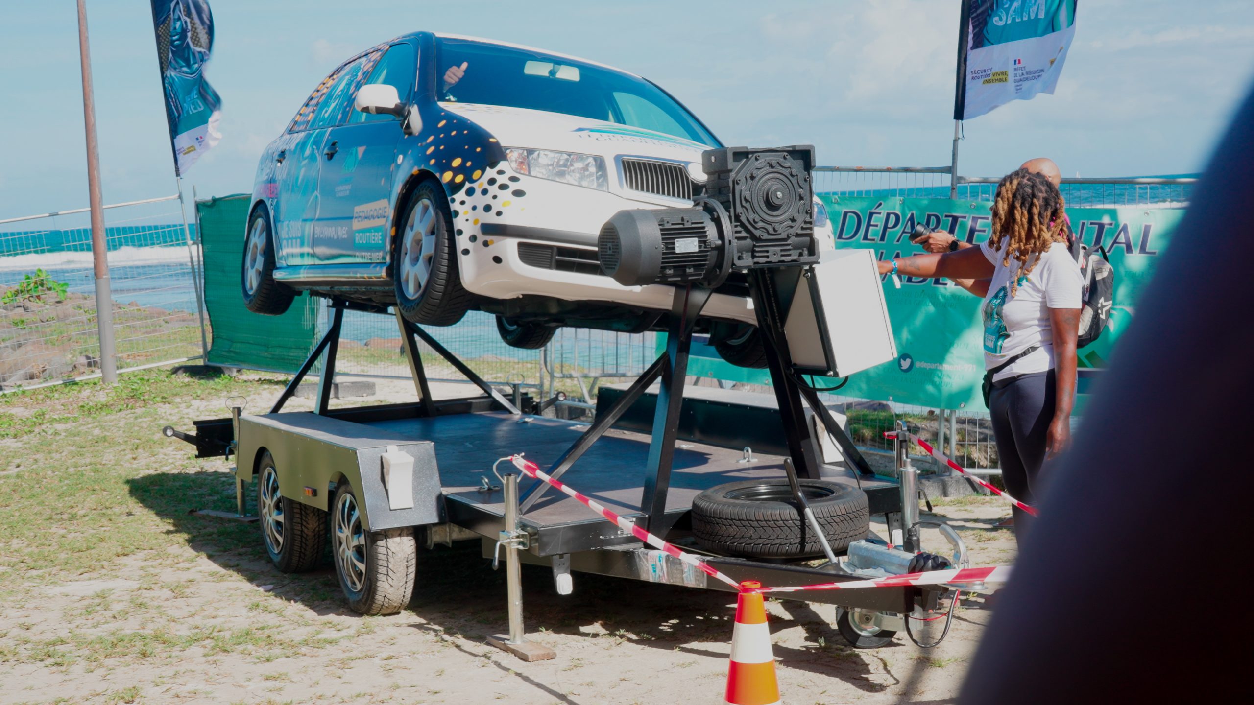 Voiture blanche montée sur un dispositif de simulation de tonneaux, entourée de barrières de sécurité et de participants à une démonstration en plein jour, face à l’océan.