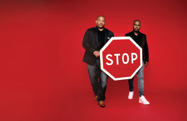Deux hommes debout sur un fond rouge vif, habillés en noir et jean, tiennent ensemble un panneau STOP rouge et blanc, symbole fort de la campagne de sensibilisation à la sécurité routière.