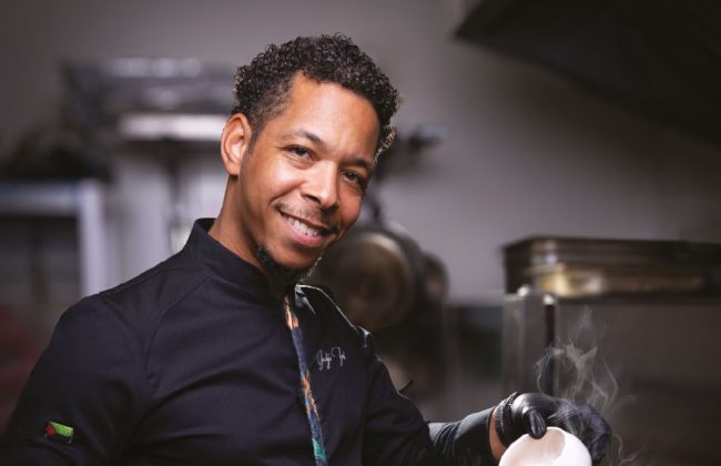 Yadji Zami, chef cuisinier vêtu de noir, dans sa cuisine avec une poile à la main fumante et qui verse une sauce à l'intérieur