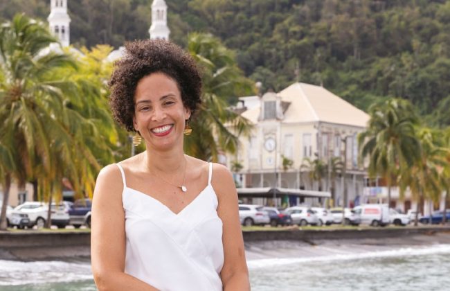 Portrait de Caroline Ventura assise sur un quai en bord de mer, souriante, vêtue d’une jupe à motifs bleus et jaunes. En arrière-plan, les bâtiments historiques et la végétation luxuriante de la commune de Saint-Pierre en Martinique.